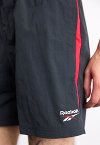 Černé sportovní šortky s červenými bočními pruhy, vyrobené z lehké tkaniny. Obsahují vyšitý logo Reebok na lemu a elastický pas.