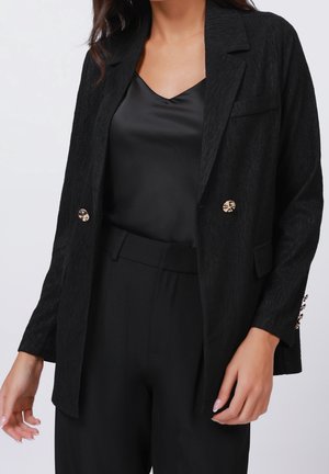 Kvinde iført en tekstureret sort blazer med gyldne knapper over en sort satin cami og sorte bukser med høj talje.