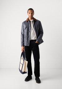 Navy jacket with brown collar, checkered shirt underneath, white t-shirt, black corduroy pants, and a canvas tote bag with blue accents.