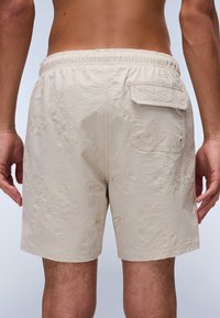 Beige zwembroek met een textuurafwerking, voorzien van een achterzak en een subtiel bloemenpatroon. Elastische tailleband voor comfort.