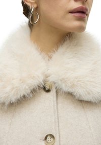 Manteau en laine beige avec un col en fausse fourrure crème, doté de grands boutons et d'une texture lisse. Gros plan sur les détails du col et de l'encolure.