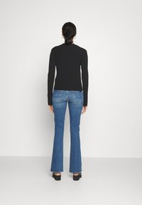 Kvinde står vendt mod en hvid væg, iført en sort langærmet top, blå bootcut-jeans og sorte platformsko.