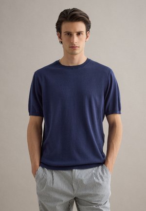 Mann trägt einen marineblauen kurzärmeligen Pullover und weiße Hosen mit dünnen vertikalen schwarzen Streifen, steht mit den Händen in den Taschen.