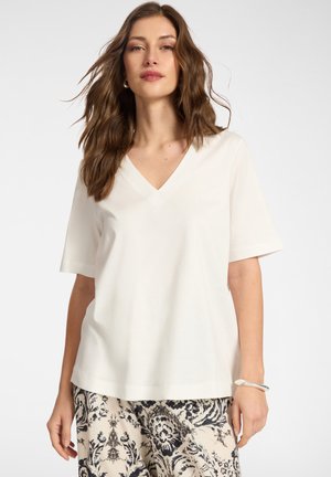 Frau mit welligem braunem Haar trägt ein schlichtes weißes V-Ausschnitt-Kurzarmshirt und gemusterte schwarz-weiße Hose, steht vor hellem Hintergrund.