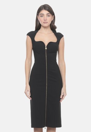 Femme portant une robe midi noire ajustée avec des manches courtes et une fermeture éclair dorée à l'avant, debout devant un fond clair uni.