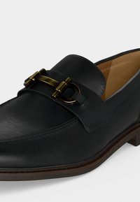 Mocassin en cuir noir avec doublure intérieure couleur fauve et détail de boucle métallique dorée sur le dessus, posé sur une surface blanche.