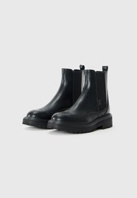 Black leather Chelsea boots with brogue detailing, elastic sides, pull tabs, and chunky rubber soles; sleek design and smooth texture.