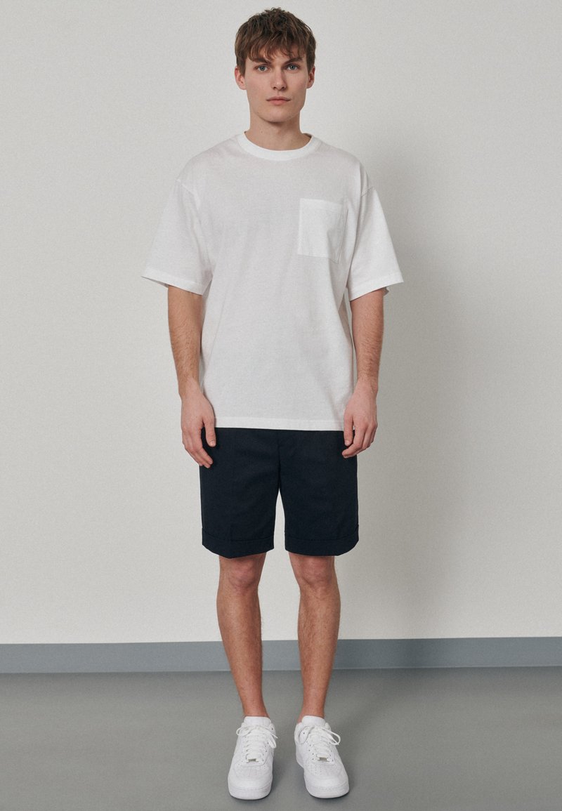 T-shirt en coton blanc avec une poche poitrine, associé à un short bleu marine à revers. Modèle portant des baskets blanches, se tenant devant un fond neutre.