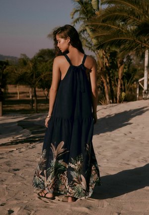 Woman in a sleeveless navy maxi dress with floral patterns and sandals standing on sandy ground near palm trees at sunset.