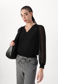 Blouse noire à manches longues avec des pois texturés, décolleté en V et taille élastique. Associée à un pantalon gris clair avec cordon de serrage et un sac noir.