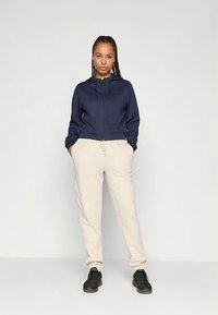 Kobieta stoi z rękami w kieszeniach, ubrana w granatowy rozpinany hoodie, beżowe dresy i czarne sneakersy na jednolitym jasnym tle.