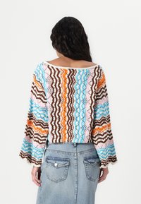 Stickad topp med off-shoulder design, med ett vågigt mönster i brunt, blått, rosa och orange. Långa trumpetärmar, lätt textur.