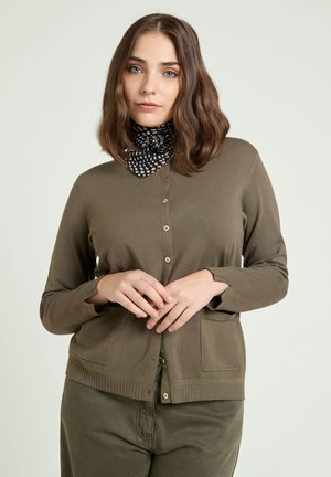 Woman with shoulder-length brown hair wears olive cardigan and pants with a black and white patterned neck scarf, hands clasped in front.