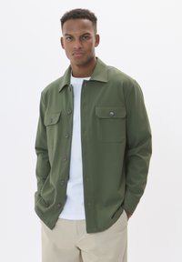 Veste verte à boutons avec deux poches poitrine, un col standard et une texture lisse, portée par-dessus un t-shirt blanc.