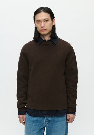 Brown knitted sweater with a ribbed texture, round neckline, and long sleeves, layered over a dark blue shirt. Light blue denim jeans.