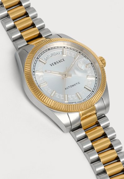Montre automatique Versace argent et or affichant samedi et 10h10 avec bracelet en maillons métalliques et lunette cannelée en or.