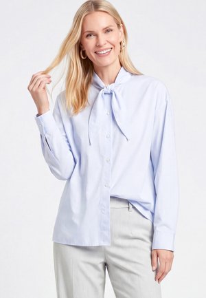 Blonde Frau lächelt, trägt eine hellblaue Bluse mit Knöpfen und Krawatte sowie hellgraue Hose, hält mit einer Hand eine Haarsträhne.