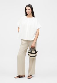 Γυναίκα με άνετο λευκό τοπ με κοντά μανίκια, μπεζ παντελόνι με φαρδιά μπατζάκια, μαύρες σαγιονάρες, κρατώντας μικρή υφαντή μαύρη και μπεζ τσάντα, σε μονόχρωμο φόντο.