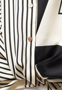 Chemise boutonnée avec rayures verticales crème et noires, tissu lisse et ourlet arrondi. Boutons marron visibles près de l'encolure.