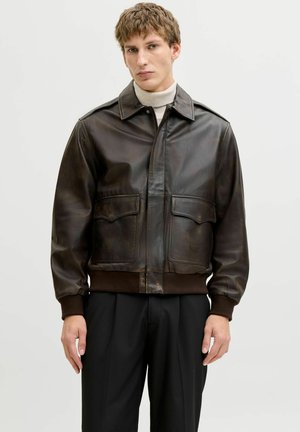 Fiatal férfi sötétbarna bőr bombázó dzsekit visel bézs garbó és fekete nadrág fölött, egy egyszerű világos háttér előtt állva.