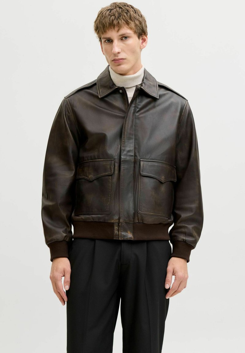 Fiatal férfi sötétbarna bőr bombázó dzsekit visel bézs garbó és fekete nadrág fölött, egy egyszerű világos háttér előtt állva.