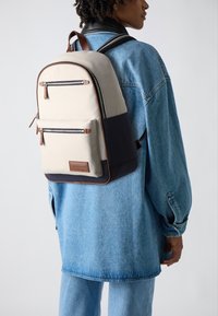 Zaino beige e blu navy con dettagli marroni, dotato di due tasche frontali con cerniera, una forma strutturata e spallacci regolabili.