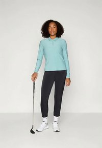 Kvinne i blå langermet polo, sorte bukser og hvite joggesko som holder en golfkølle og står mot en enkel lys bakgrunn.