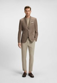 Brunrutigt kostymjacka med slag, kombinerad med en beige skjorta, ljusbeiga byxor och mörkbruna loafers.