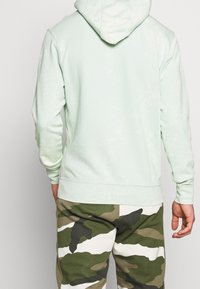 Persona che indossa una felpa leggera color verde menta e pantaloni camouflage nei colori verde, bianco e marrone, in piedi di spalle rispetto alla macchina fotografica.