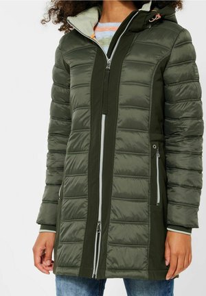 Femme portant une veste d'hiver matelassée olive mi-longue avec fermeture éclair à l'avant et poches latérales zippées.