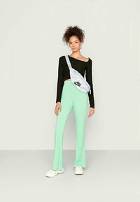 Top cropped de manga longa preto, calças fluídas verdes claras e uma bolsa de cintura cinza com um logótipo preto. O modelo usa ténis brancos.
