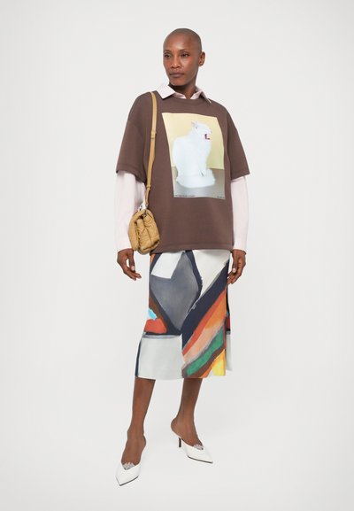 Brązowy, oversize'owy t-shirt z grafiką białego królika, nałożony na jasnoróżową bluzkę z długim rękawem, w połączeniu z kolorową spódnicą w wzory i białymi obcasami.