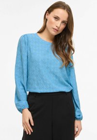 Hellblaue langärmelige Bluse mit floraler Lochstickerei, rundem Ausschnitt und elastischen Bündchen, kombiniert mit hoch taillierter schwarzer Hose.