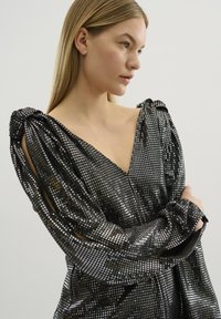 Top nero con paillettes, scollatura a V profonda, maniche a sbuffo e dettagli a taglio. Il tessuto brilla con un motivo testurizzato a griglia.