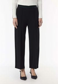 Pantaloni neri a gamba larga con una texture liscia, dotati di una vita scura con scritte in rilievo e piccole aperture laterali all'orlo.