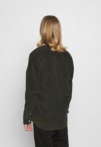 Person med skulderlangt blond hår iført en mørkegrøn kordfløjlsskjorte og sorte bukser, vendt væk mod en ensfarvet lys baggrund.