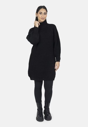 Kvinde i sort ribbet rullekrave-strikket kjole, sorte leggings og snørebånds chunky sorte støvler, poserer med den ene hånd nær ansigtet.