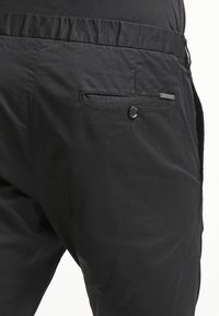 Svarta tygbyxor med elastisk midja, försedd med en sidoficka med knappstängning och en liten varumärkesetikett på baksidan.