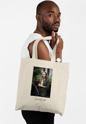 Mężczyzna z brodą i zegarkiem trzyma beżową torbę na ramię z wizerunkiem kobiety w zielonym stroju trzymającej czerwoną teczkę, pod spodem napis „gossip girl”.