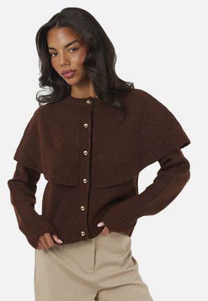 Vrouw met lang donker haar die een bruine cardigan met knopen draagt met een brede geribbelde cape en beige broek tegen een witte achtergrond.