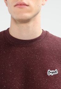Detailní záběr na osobu v bordó svetr s tečkami, která má na hrudi malý bílý nášivku "Originals".