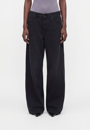 Personne portant un jean noir à jambes larges et des chaussures noires pointues, debout devant un fond clair uni.