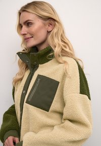 Giacca in pile beige e verde, con colletto alto, tasca sul petto con zip e dettagli in tessuto testurizzato. Bottoni lungo la parte anteriore.