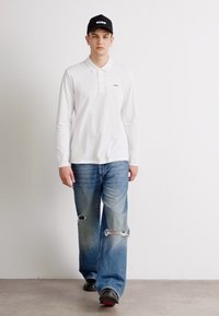 Hvit langermet polo skjorte med krage, sammen med lyseblå slitte jeans med store hull. Svart caps med "HUGO" logo.