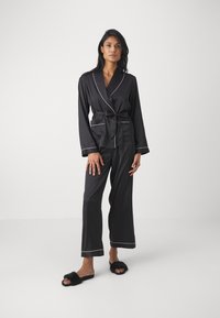 Svarta pyjamas med satinfinish, med en sjalkrage, långa ärmar och kontrasterande vita kantband. Modellerna bär svarta fluffiga tofflor.