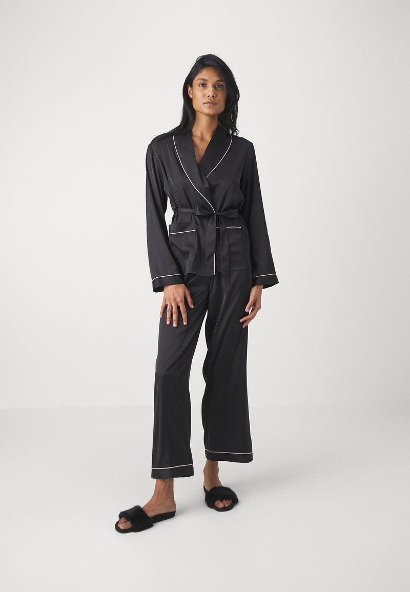 Svarta pyjamas med satinfinish, med en sjalkrage, långa ärmar och kontrasterande vita kantband. Modellerna bär svarta fluffiga tofflor.
