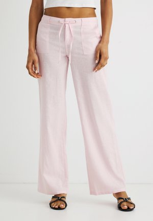 Femeie purtând pantaloni largi din in, roz deschis, cu buzunare aplicate în față și talie cu șnur, asortați cu sandale negre cu degetele descoperite.