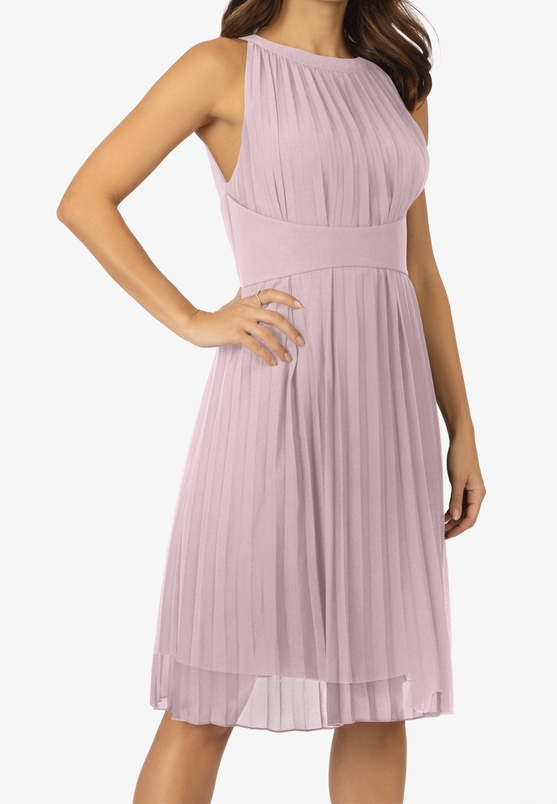 Robe midi plissée sans manches violet clair avec une taille cintrée, portée par une femme debout avec une main sur la hanche.