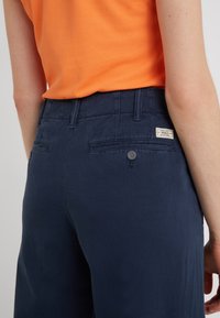 Mörkblå byxor med en slät textur, med synlig knapp och fickdetaljer, tillsammans med en orange skjorta. Poloetikett på linningen.