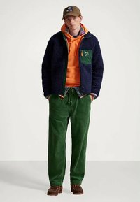 Νεαρός άνδρας με καφέ καπέλο, μπλε φλις μπουφάν πάνω από πορτοκαλί φούτερ και καρό πουκάμισο, με πράσινα βελούδινα παντελόνια και καφέ παπούτσια.